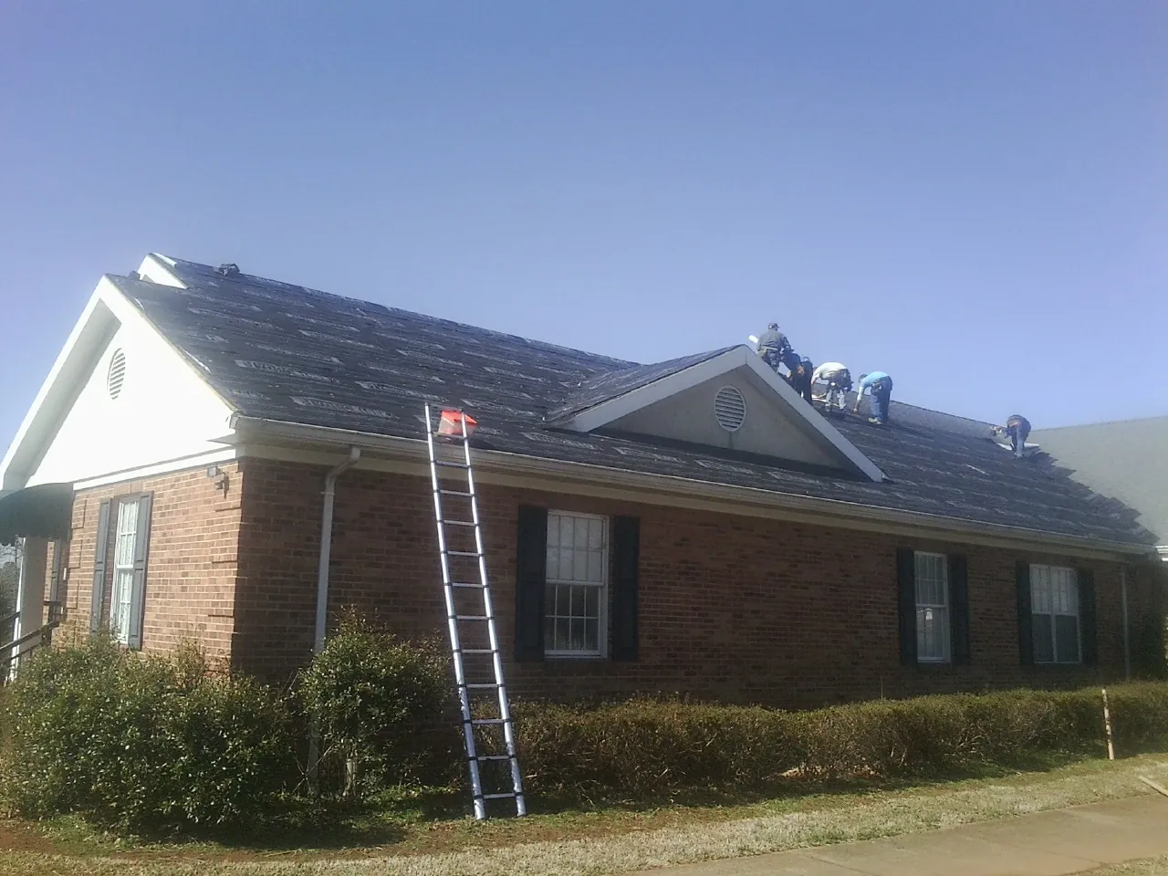 Crew tearing shingles off a brick ranch roof with a ladder set — Metal Roof Repair work by our crew in Rainsville