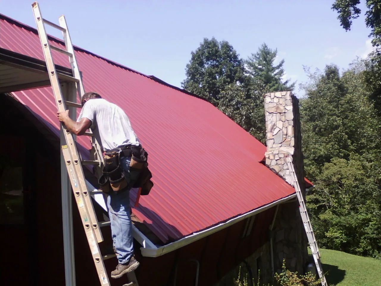 Roofer on a ladder beside a red metal roof and stone chimney — Soffit & Fascia Repair by Roofing Brothers in Rainsville